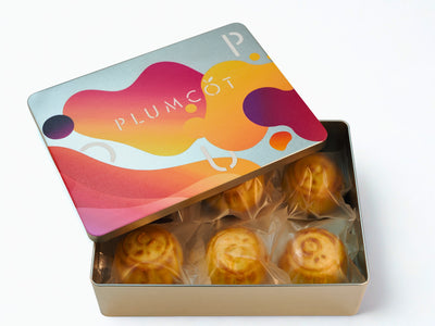 Colorful tin box with 'PLUMCOT' branding containing individually wrapped mooncakes.